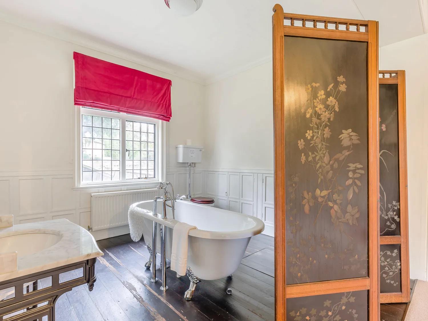 Elegant bathroom with a freestanding clawfoot bathtub, decorative folding screen, marble sink, and a window with a red Roman shade.