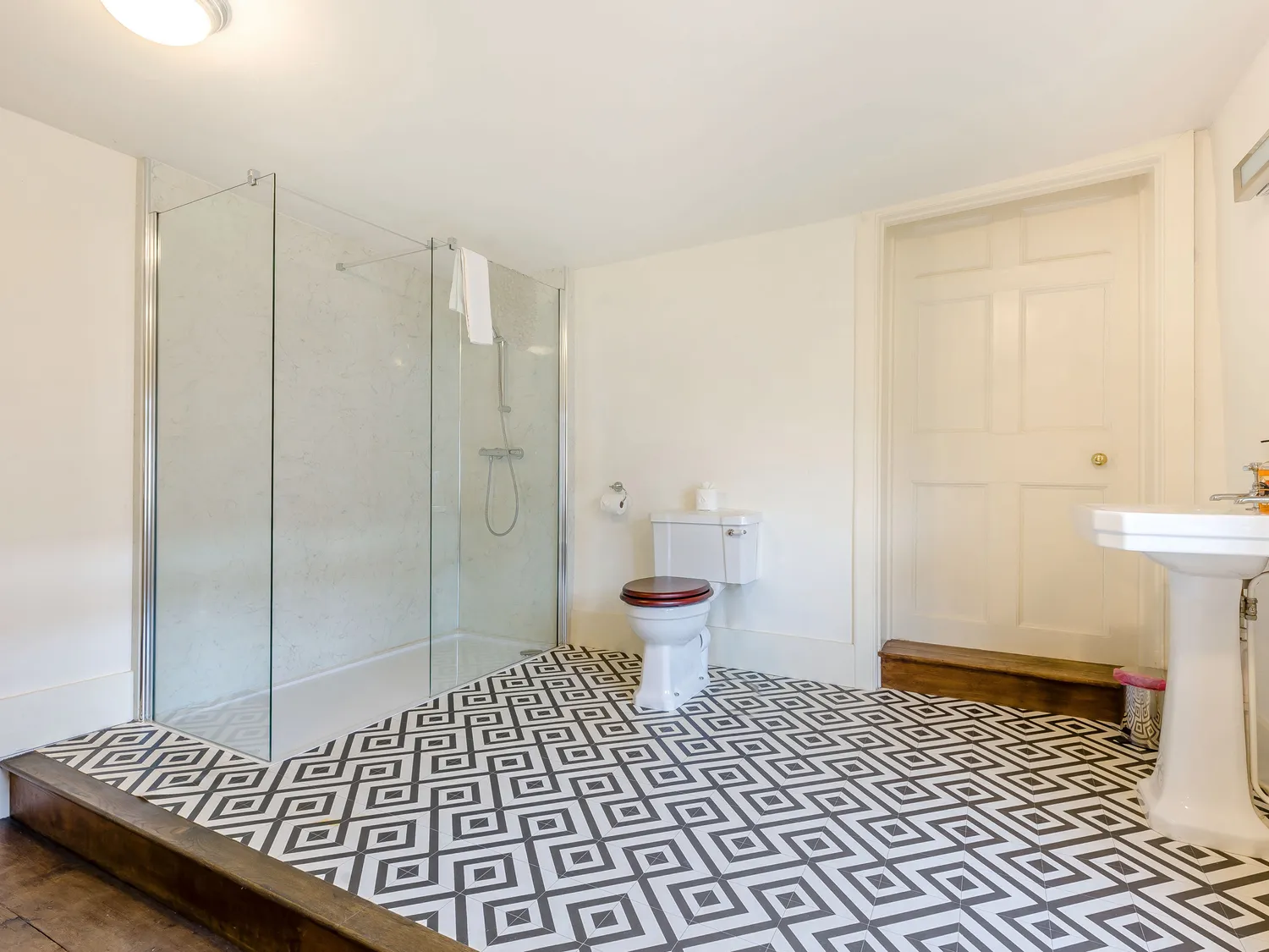 Modern bathroom with geometric tile flooring, glass shower enclosure, white toilet, and pedestal sink.