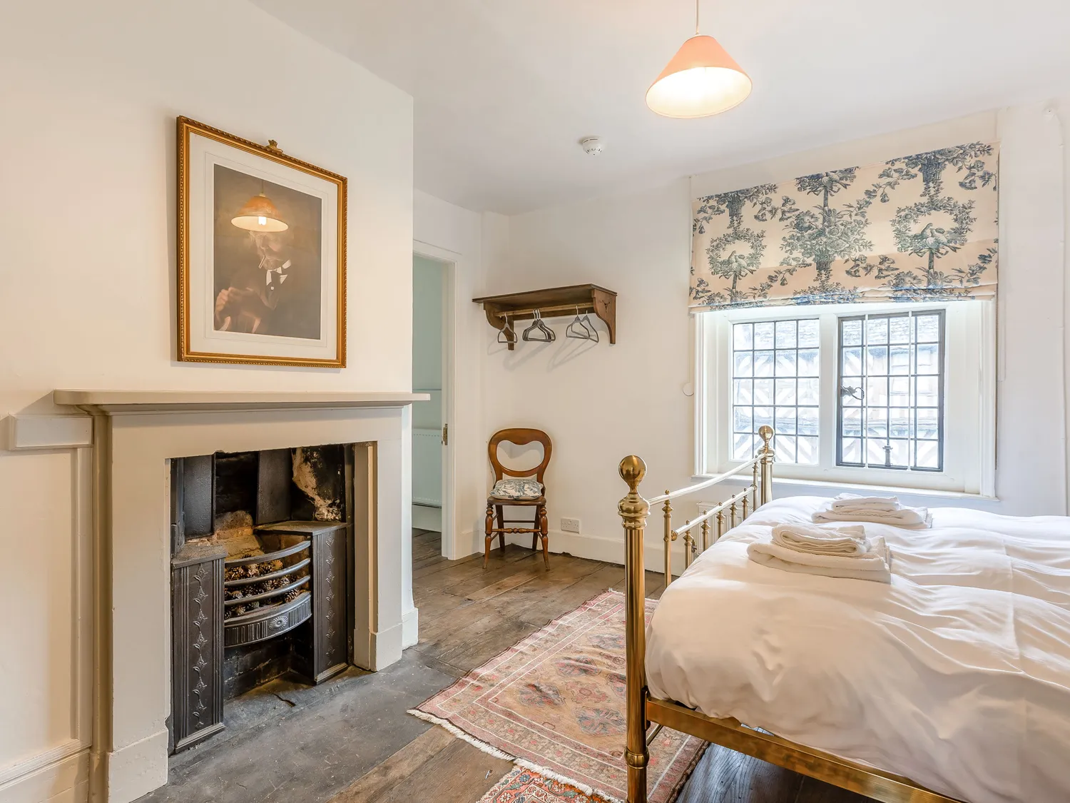 Cozy vintage bedroom with brass bed, fireplace, framed artwork, wooden floor, and floral window shade.