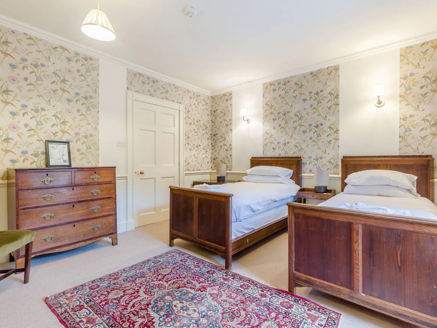 Vintage bedroom with twin wooden beds, floral wallpaper, antique dresser, and ornate rug.