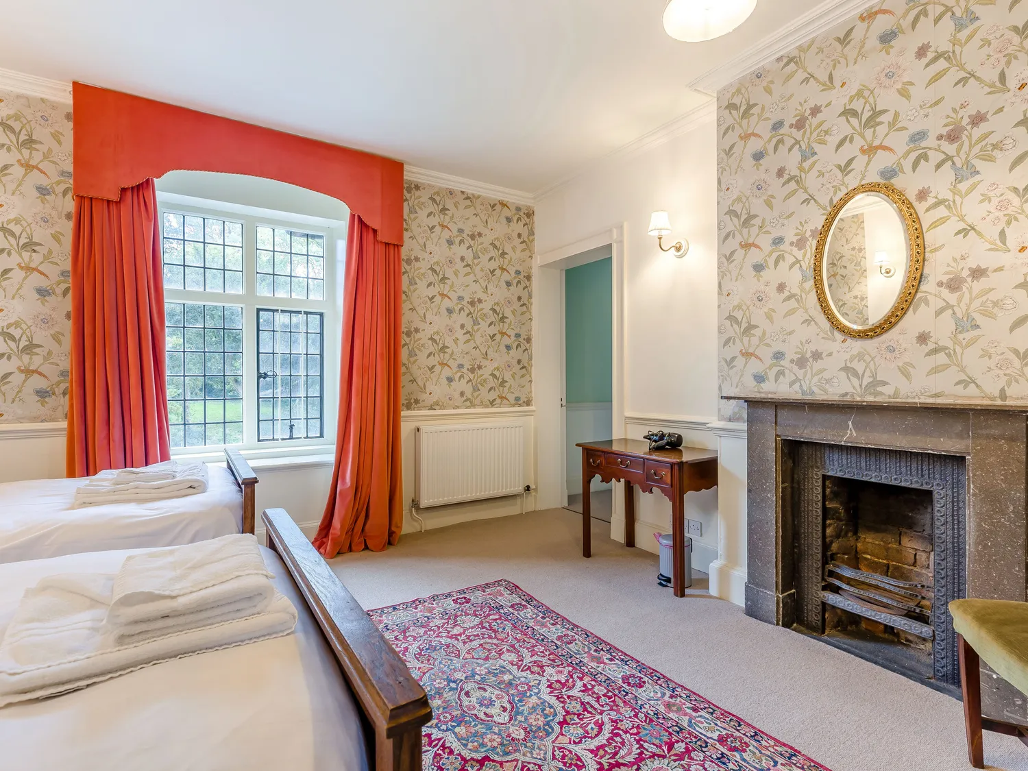 Cozy bedroom with twin beds, vintage fireplace, floral wallpaper, red curtains, and large window with garden view.