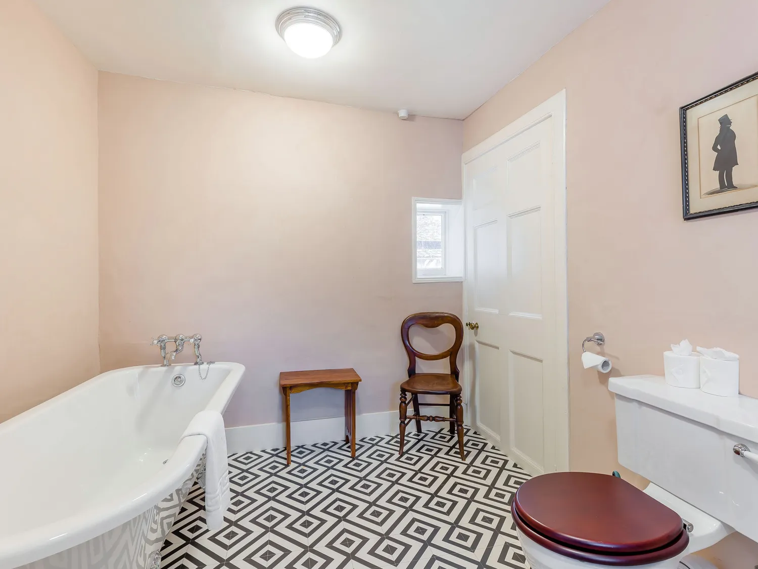 Vintage bathroom with clawfoot tub, patterned tile floor, wooden chair, and wall art.