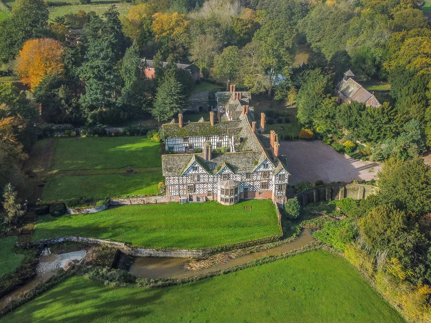 Aerial view of a historic Tudor-style mansion surrounded by lush greenery and trees, with a small stream flowing nearby.