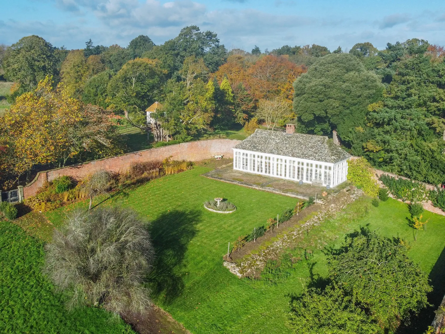 Aerial view of a historic greenhouse surrounded by lush green gardens and mature trees in a scenic countryside setting.