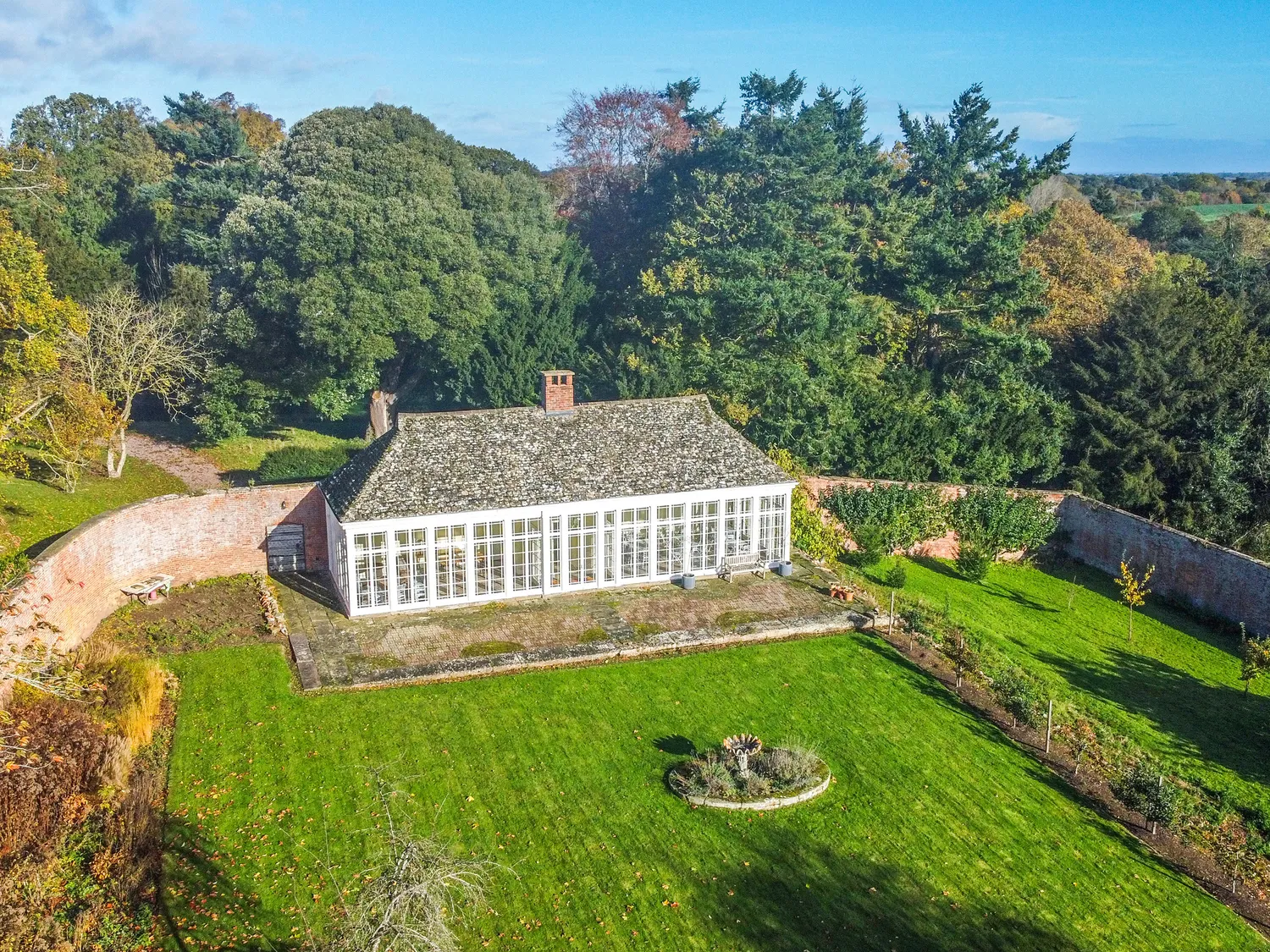 Aerial view of a charming brick house with a large garden, surrounded by lush greenery and trees, under a clear blue sky.