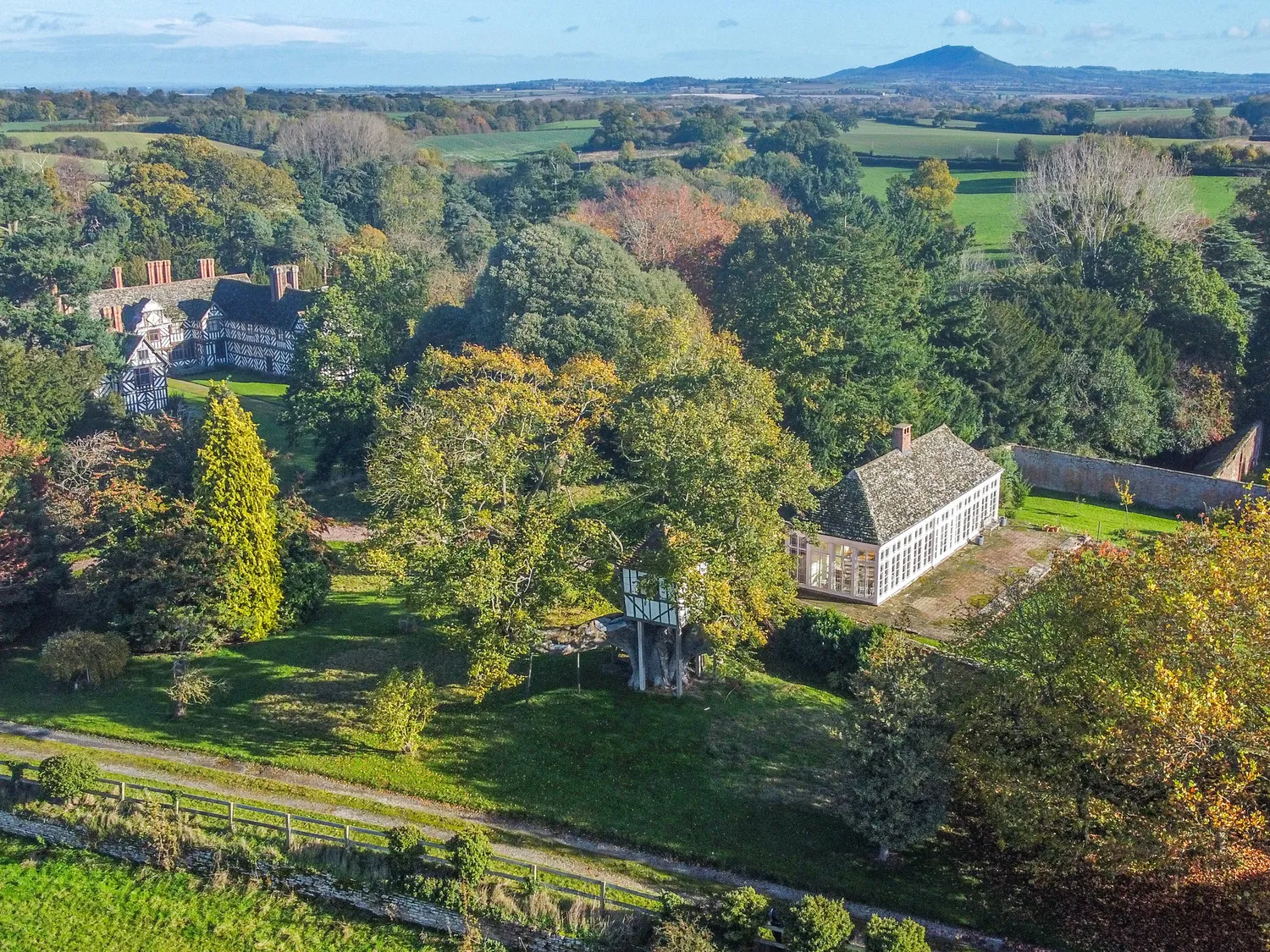 Aerial view of a historic estate with half-timbered buildings surrounded by lush greenery and trees, set against a backdrop of rolling hills and a distant mountain.