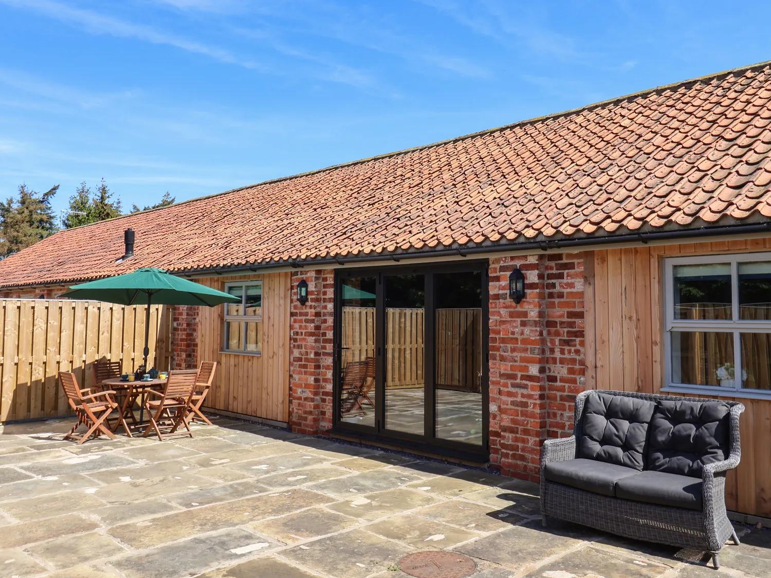 Charming brick and wood cottage with tiled roof, featuring a spacious patio with outdoor seating and a green umbrella.