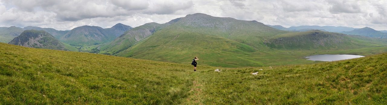 Panoramic view of lush green hills and mountains with a hiker walking along a path, under a cloudy sky, showcasing the natural beauty of the landscape.