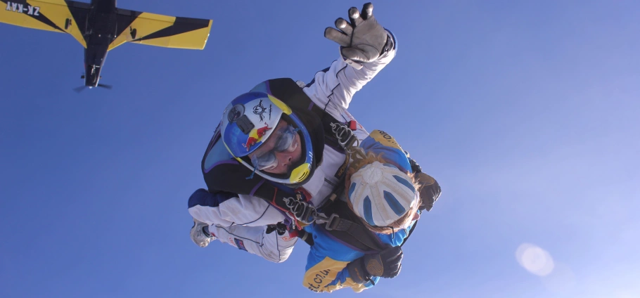 Two people skydiving with parachutes, blue sky background, airplane in the distance.