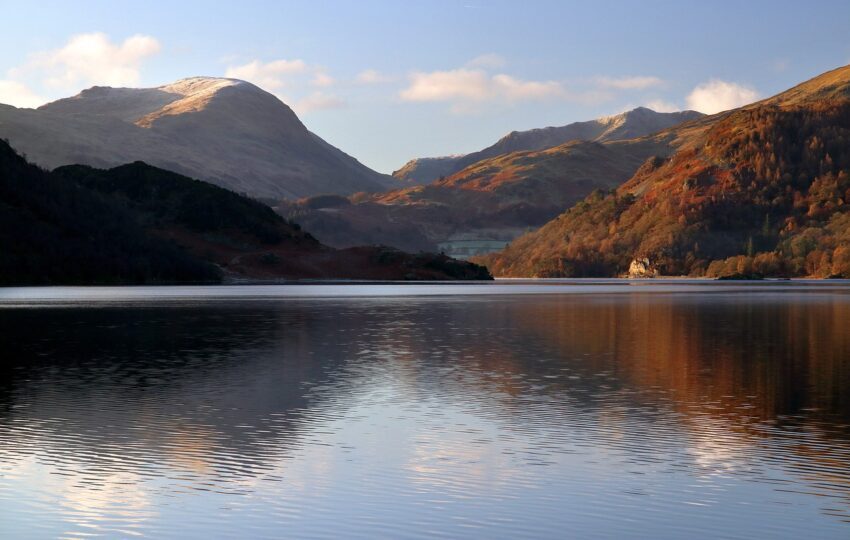 Cumbria | Scenic view of a tranquil lake surrounded by mountains, featuring autumn foliage and a clear blue sky, with reflections of the landscape on the water's surface.