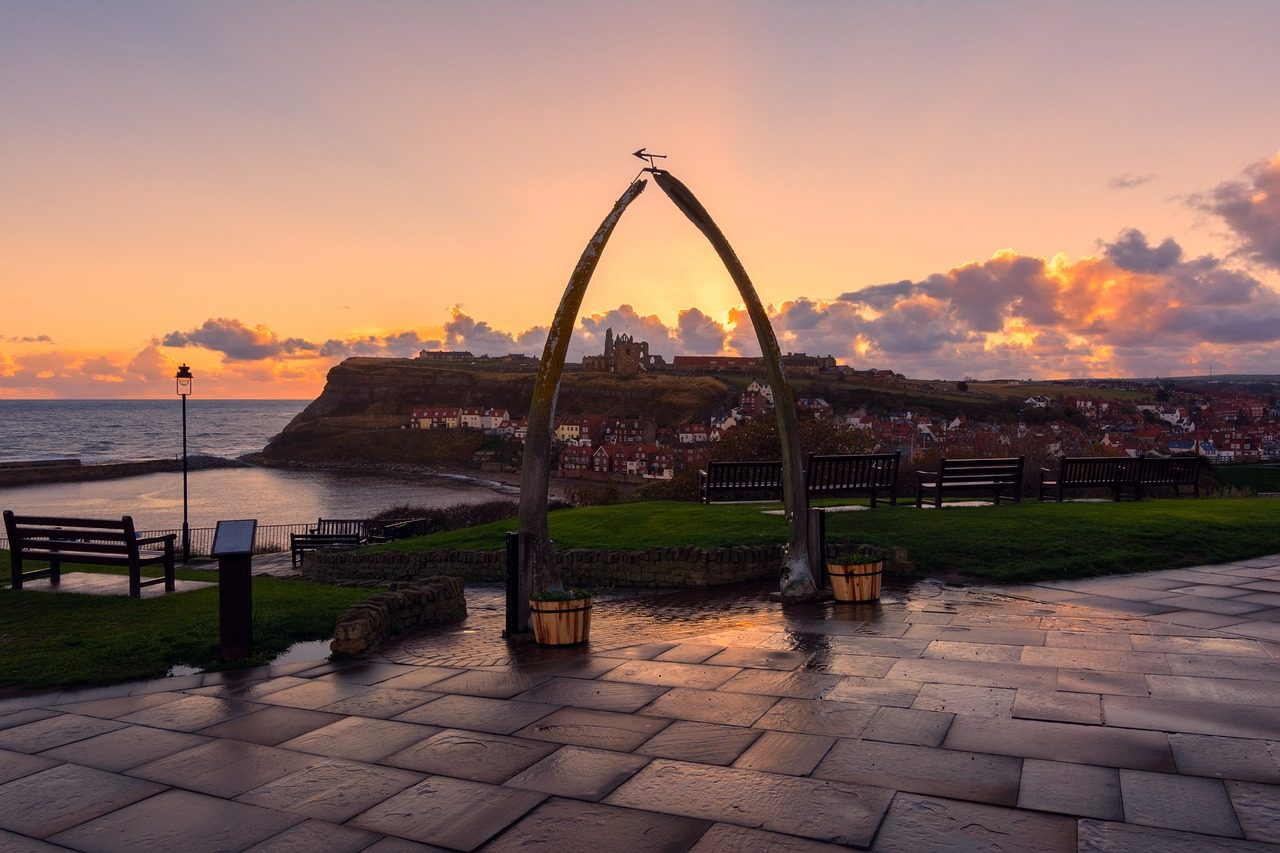 View of a whale bone arch at sunset overlooking the coastal town of Whitby, with colorful clouds and the ruins of Whitby Abbey in the background, showcasing the scenic beauty of the Yorkshire coastline.