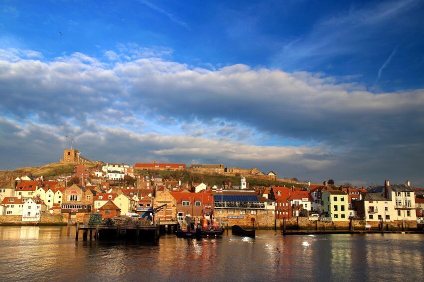 Coastal view of Whitby, England, featuring colorful houses along the harbor, a historic church on the hill, and dramatic clouds in the sky.