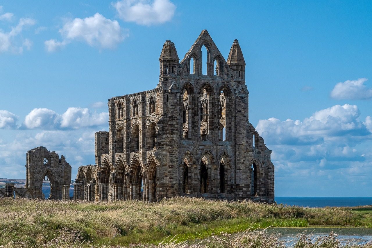 Ruins of Whitby Abbey under a bright blue sky, surrounded by lush grass and fields, with the North Sea in the background. The historic stone structure features Gothic architectural elements, including pointed arches and large windows, showcasing its medieval heritage.