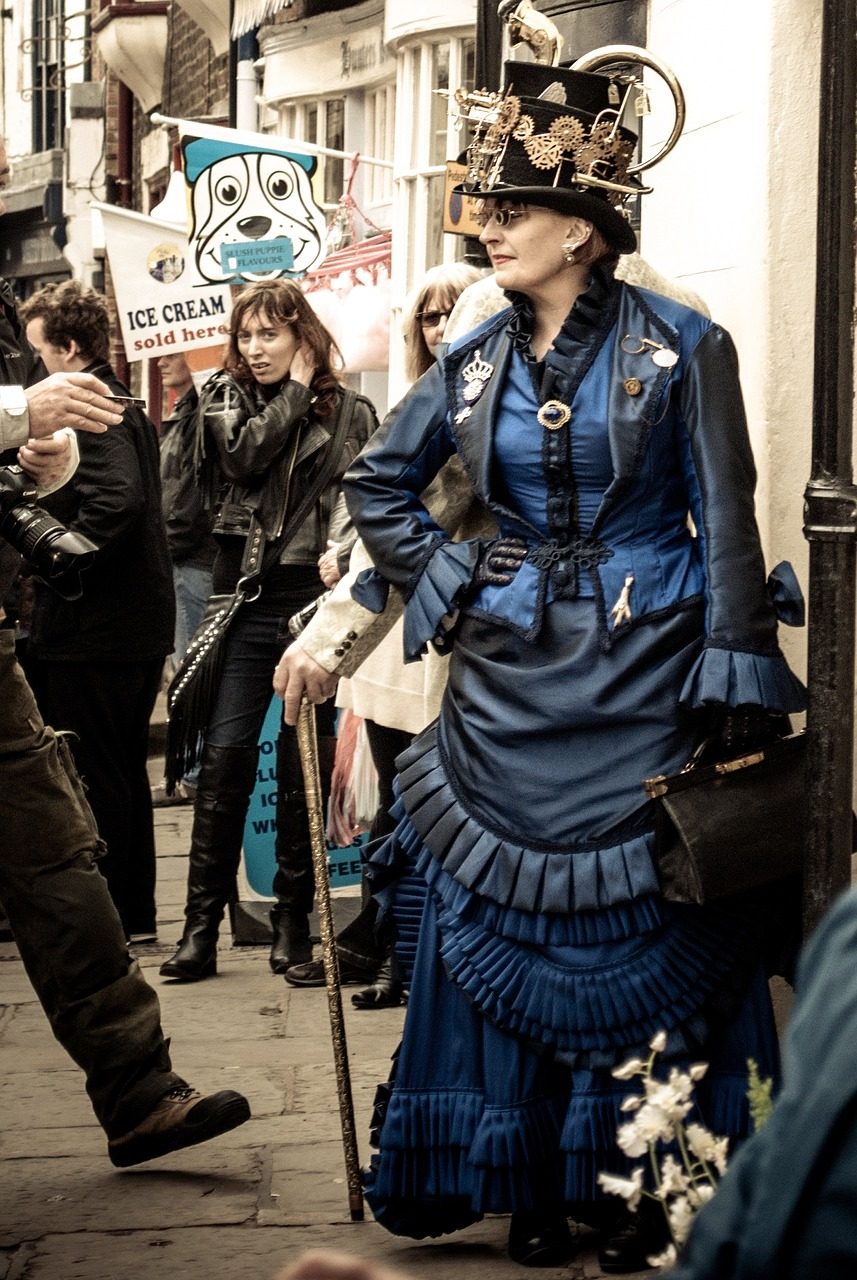 A woman in a vintage blue dress with ruffles and a unique hat stands on a street, surrounded by onlookers. She holds a cane and carries a small handbag, while a sign advertising ice cream flavors is visible in the background.