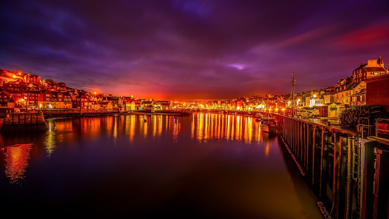 Night view of a picturesque harbor with colorful illuminated buildings reflecting on calm water, under a dramatic sky in a coastal town.