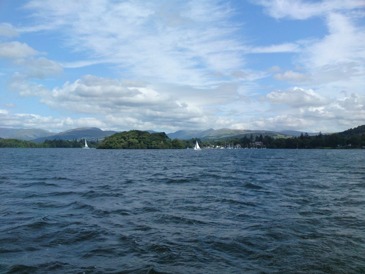 Scenic view of a tranquil lake surrounded by lush greenery and distant mountains, featuring several sailboats on the water under a partly cloudy sky. Ideal for nature lovers and sailing enthusiasts.